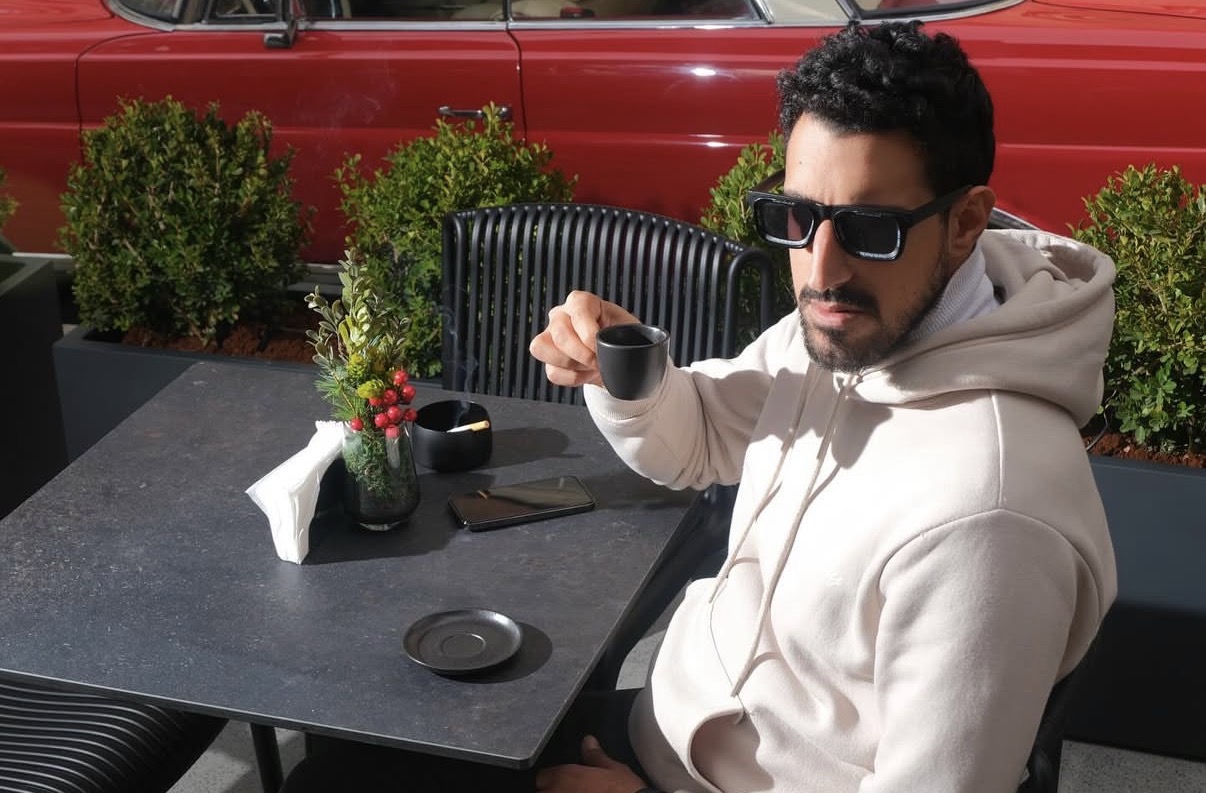Malek in sunglasses with an espresso in front of a red vintage car